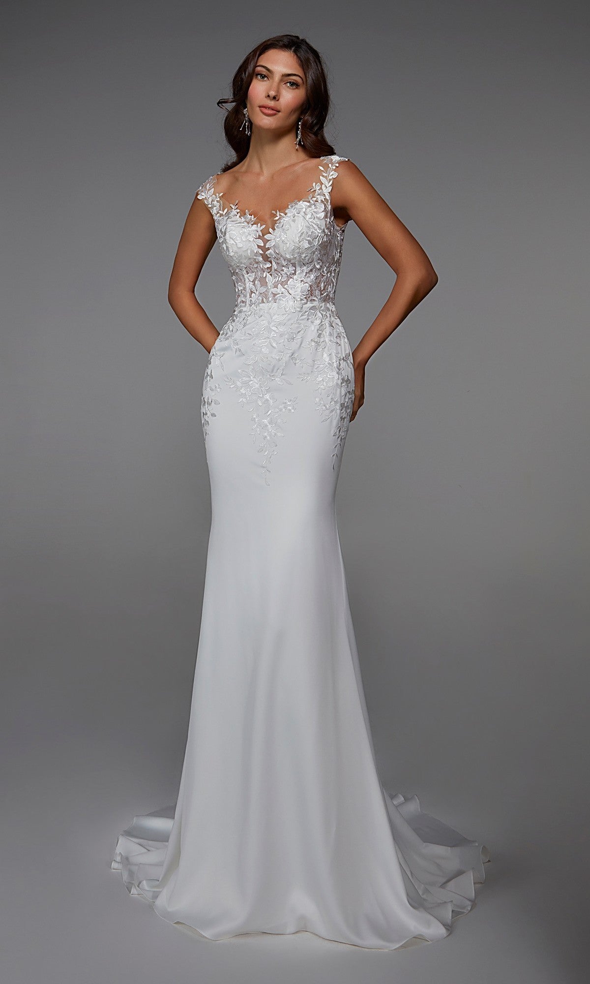 A woman wearing the Alyce 7026 Long White Dress in Diamond White, a sleeveless gown with intricate lace bodice detailing and a fitted, floor-length skirt, stands against a plain gray background.